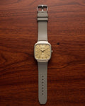 A Rado watch with a square beige dial and a light gray strap, laid flat on a dark brown wooden surface.
